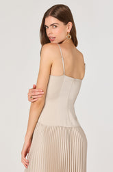 A woman is wearing a sleeveless, beige dress with thin spaghetti straps. The upper part of the dress is smooth and fitted, while the lower part features pleated fabric. She has wavy brown hair, large circular earrings, and a ring on her right hand.
